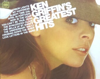Op de afbeelding: Een vintage vinyl platenhoes met een vrouw die een bruine cowboyhoed draagt. De albumtitel is "Ken Griffin's Greatest Hits" en vermeldt de nummers "Marbor Lights", "My Happiness", "Sleepy Time Gal", "April in Portugal", "Now is the Hour", "You Can't Be True, Dear", "The Syncopated Clock", "The Bells of St. Mary's", "The Anniversary Waltz", "Cruising Down the River" en "Love Letters in the Sand".