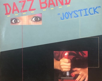 Puede incluir: Una portada de disco de vinilo para el álbum "Joystick" de la banda Dazz Band. La portada presenta los ojos de una mujer y un joystick rojo.