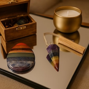 May include: A wooden box, a gold candle holder with a white candle, and a mirrored surface. A rainbow-colored stone and a crystal pendulum with a wooden base are also present. The pendulum is suspended by a silver chain.