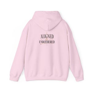 May include: A light pink hoodie with the words "ALIGNED & UNBOTHERED" printed in a dark brown script on the back. The hoodie has a hood and long sleeves.