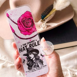May include: A white bookmark with a pink rose and a cowboy hat design. The text on the bookmark reads "The Cowboy Hat Rule" and "You wear the hat, you ride the cowboy, Chestnut Springs".