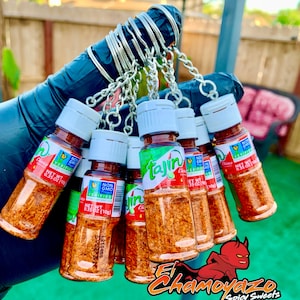 May include: A set of seven small clear plastic bottles with red lids and red labels. The bottles are filled with a red and orange spice blend. The bottles are attached to a silver key chain. The keychain is labeled "El Chamoyazo Spicy Sweets".