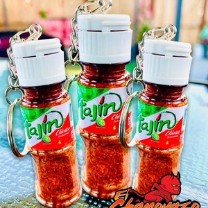 May include: Three miniature glass bottles with red and green labels and white lids. The labels say "Tajin Clasico Seasoning" with a lime image. The bottles are filled with red chili powder and attached to key rings. The bottles are on a blue and white surface with the text "El Chamoyazo Spicy Sweets".