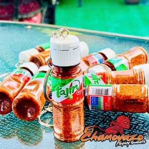 May include: A group of small, clear plastic bottles filled with a red and orange powder. The bottles have a white lid and a silver ring. The bottles are labeled "Tajin" and have a green lime image. The bottles are on a green and blue surface. The text "El Chamoyazo Spicy Sweets" is visible in the bottom right corner of the image.
