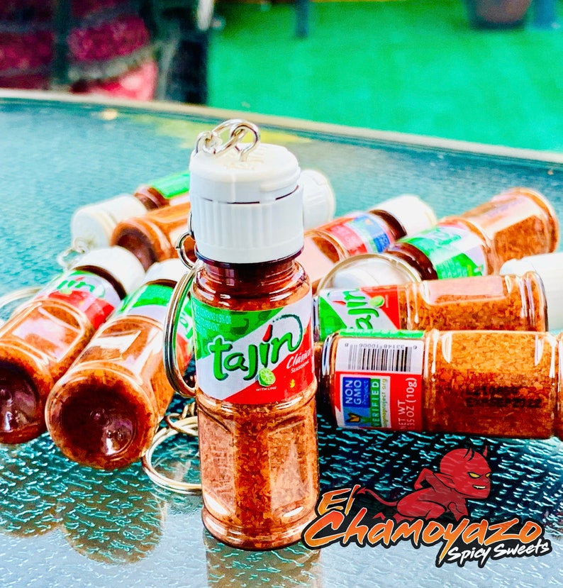 May include: A group of small, clear plastic bottles filled with a red and orange powder. The bottles have a white lid and a silver ring. The bottles are labeled "Tajin" and have a green lime image. The bottles are on a green and blue surface. The text "El Chamoyazo Spicy Sweets" is visible in the bottom right corner of the image.