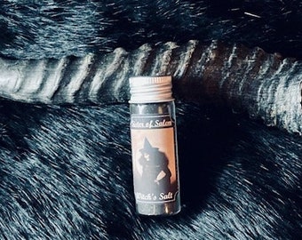 May include: A small glass bottle with a silver lid containing black salt. The bottle has a label that reads "Witches of Salem" and "Witch's Salt". The bottle is sitting on a black fur surface.