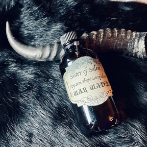 May include: A dark glass bottle with a black label that reads "Sister of Salem" and "War Water" with a website address. The bottle is sitting on a black fur surface.