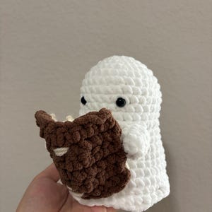 May include: A white crocheted ghost figurine with small black eyes, holding a small brown crocheted book with visible white pages. The ghost has a rounded head and a soft, textured yarn surface, appearing as a handmade plush toy item.