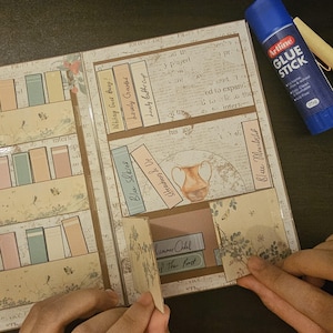 Puede incluir: Un álbum de recortes hecho a mano con un diseño de estantería, con títulos de libros en miniatura como "Blue Skies" y "Summer Child of The Forst". Un pegamento en barra Artline azul es visible. El álbum tiene una estética vintage.