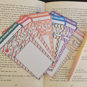 May include: A selection of reading challenge bookmarks in various colours and patterns, including stripes. Each bookmark has space for time, guidelines, and audio speed. A pen rests on an open book.