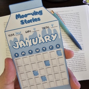 May include: A blue and white paper calendar shaped like a milk carton, with the text "Moo-ving Stories" at the top. The calendar is for January 2025, with a goal of 4. A blue and green pen rests on the open book.