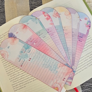 Op de afbeelding: Een verzameling bloemrijke bladwijzers uitgespreid over een open boek. Elke bladwijzer heeft een aquarelontwerp met ruimte voor titel, genre en pagina-informatie. Het kleurenpalet omvat roze, blauw en paars.