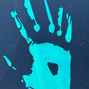 May include: A teal handprint sticker on a dark blue surface. The handprint is a detailed outline of a hand, with visible fingers and palm. The teal color contrasts with the dark background, creating a striking visual effect.