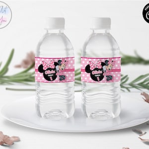 K&ouml;nnte beinhalten: Zwei transparente Wasserflaschen mit wei&szlig;en Verschl&uuml;ssen, jeweils mit einem rosa Etikett mit einem Minnie-Mouse-Design versehen. Die Etiketten zeigen einen schwarzen Minnie-Mouse-Kopf mit einer rosa Schleife, den Namen "Olivia" und die Zahl "1".
