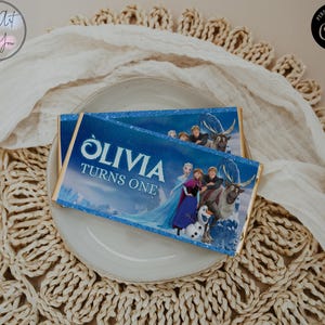 Puede incluir: Dos envoltorios azules de barras de chocolate con el texto "OLIVIA TURNS ONE" e imágenes de personajes de la película Frozen. Los envoltorios están sobre un plato blanco, sobre un salvamanteles tejido y un paño blanco.
