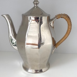 Könnte beinhalten: Eine silberne Teekanne mit einem geflochtenen Weidenhenkel. Die Teekanne hat einen runden Körper mit einem facettierten Design.