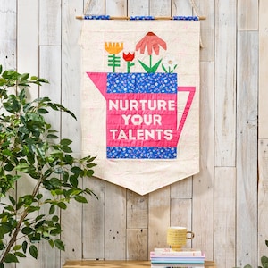 May include: A fabric wall hanging featuring a quilted watering can design in pink and blue floral patterns. The text "NURTURE YOUR TALENTS" is printed in white block letters on the center of the watering can, which is topped with colorful fabric flowers.