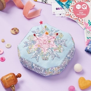 May include: A blue and white floral pincushion with a star-shaped design and pink accents. The pincushion is filled with pins and is sitting on a purple surface. The image also includes a wooden spool of thread, a roll of pink ribbon, and a variety of fabric scraps.