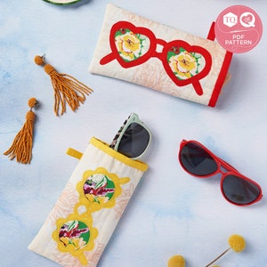 Könnte beinhalten: Zwei Stoff-Brillenetuis mit Blumenmustern. Ein Etui ist rot mit einem herzförmigen Design und das andere ist gelb mit einem runden Design. Die Etuis befinden sich auf einem hellblauen Hintergrund mit einer roten Sonnenbrille und einer grünen Sonnenbrille.