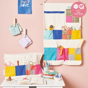 Könnte beinhalten: Ein weißer gesteppter Wandorganizer mit bunten Stofftaschen. Der Organizer hat verschiedene Taschen in unterschiedlichen Größen und Formen, perfekt zum Verstauen von Nähzubehör. Der Organizer hängt an einer rosa Wand mit einem Schild, auf dem "Just Love" steht.