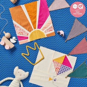 May include: Two fabric quilt blocks, one with a sunburst design in orange, yellow, pink, and white, and the other with a kite design in yellow, pink, purple, and white. Both blocks are on a blue background with white stars.