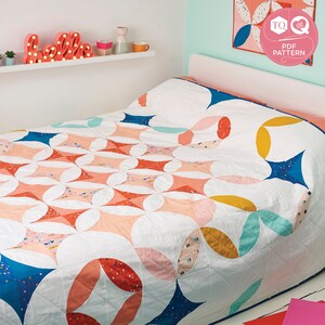 May include: A white, pink, blue, and yellow quilt with a geometric pattern. The quilt is on a bed with a white headboard and a white shelf above the headboard. The shelf has a pink neon sign that says "hello" and a few decorative items.