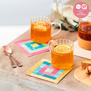 May include: Two colourful quilted coasters with a glass of lemon tea on top of each. The coasters are made of fabric squares sewn together in a patchwork pattern. The tea is in a clear glass mug with a handle. The background is a wooden table with a glass jug of tea and a plate of biscotti.