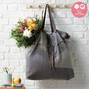 May include: A gray and white fabric tote bag with brown leather straps and a tie closure. The bag is hanging on a white brick wall with a bouquet of flowers next to it. The bag has a label that says "TQ Love PDF Pattern".