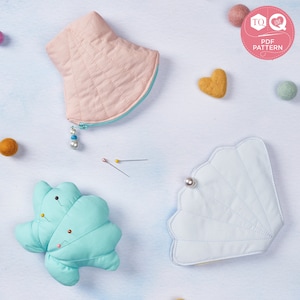 May include: Two blue and pink fabric seashell-shaped pouches with pearl accents. A smaller blue seashell-shaped pincushion with pins is in the centre of the image. The image also includes a yellow heart and several small balls of yarn.
