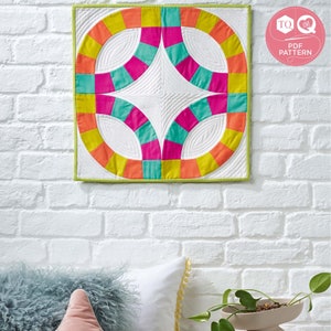 May include: A colorful quilt block with a white background and a geometric design. The quilt block is made up of orange, yellow, pink, and teal fabric. The quilt block is hanging on a white brick wall.