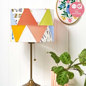 May include: A table lamp with a colourful patchwork lampshade featuring geometric patterns in shades of blue, yellow, green, and pink. The lampshade is made of fabric and has a white background. The lamp has a gold metal base and a chain pull switch.