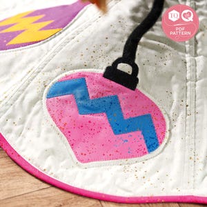 May include: A pink and blue Christmas ornament with a zig-zag pattern, sewn onto a white fabric background with gold glitter. The ornament is attached to a black string.