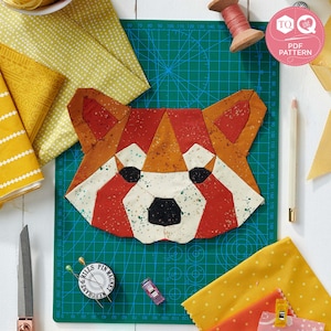 Op de afbeelding: Een geometrisch patchworkontwerp van een rode panda-gezicht, met oranje, witte en zwarte stoffen stukken. Het ontwerp ligt op een groene snijmat, omringd door stofresten, naaigerei en een klosje garen. De tekst "PDF PATTERN" is inbegrepen.