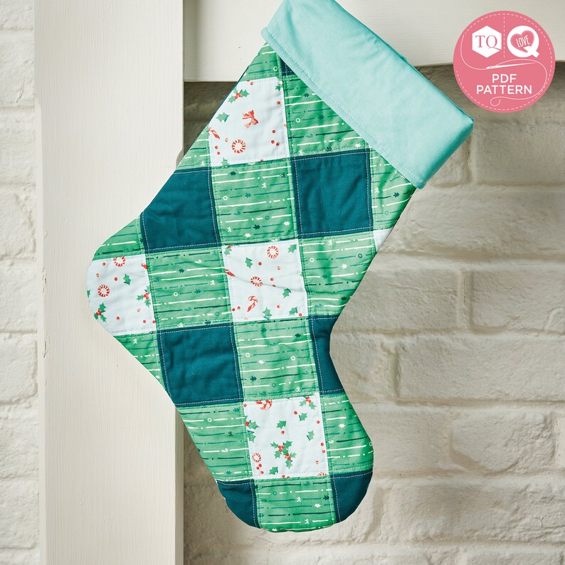 May include: A green and blue patchwork Christmas stocking with a white and green patterned fabric. The stocking has a light blue cuff and is hanging on a white wall.