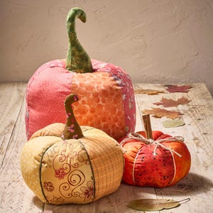 May include: Three fabric pumpkins in orange, pink, and yellow. The largest has a green stem and floral patterns. The smallest is red with a cinnamon stick and twine. Autumn-themed home decor.