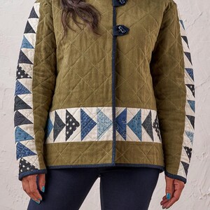 Puede incluir: Chaqueta acolchada verde oliva con detalles de patchwork geom&eacute;trico en tonos azules, blancos y negros. Presenta un patr&oacute;n de diamantes, ribete azul oscuro y cierres de palanca. Una prenda de abrigo elegante.