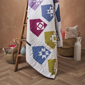 Puede incluir: Un edredón blanco con formas geométricas en morado, azul, verde y magenta. El edredón está drapeado sobre una escalera de madera. Las formas incluyen estrellas y patrones de diamantes. El edredón tiene un borde gris y se exhibe en una habitación con una combinación de colores neutros.