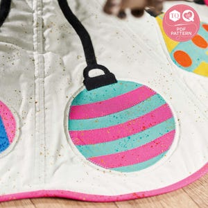 May include: A white Christmas tree skirt with a pink border and gold glitter. The skirt features three colorful ornaments with pink, blue, and green stripes.
