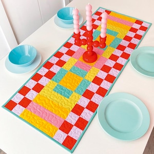 May include: A rectangular quilted table runner featuring a vibrant checkerboard pattern in red, light pink, yellow, and teal. The fabric displays a wavy, meandering stitched texture throughout, finished with a solid teal border trim.