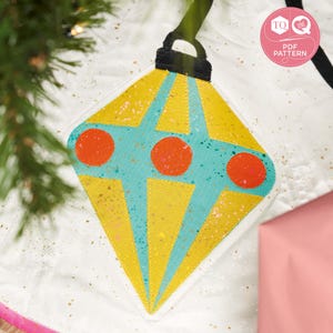May include: A white fabric Christmas tree skirt with a yellow, turquoise, and red felt ornament in the center. The ornament is shaped like a diamond and has a black felt loop at the top.