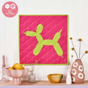 May include: A pink quilted wall hanging with a lime green balloon dog design. The square artwork has a lime green border and is displayed above a wooden shelf with decorative items, including a pink bowl of lemons and a candle.