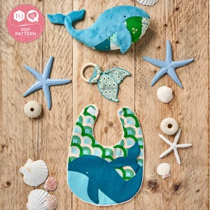 May include: A collection of baby items with a nautical theme, including a blue whale plush toy, a bib with a whale design, a whale-shaped teether, starfish, and seashells. The items are arranged on a wooden surface.