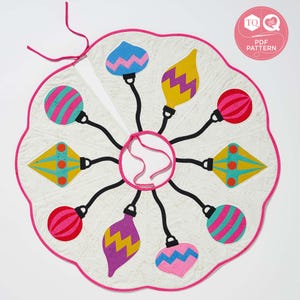 May include: A white fabric Christmas tree skirt with a pink scalloped edge. The skirt features eight colorful ornaments with different patterns and designs. The ornaments are attached to the center of the skirt with black strings.