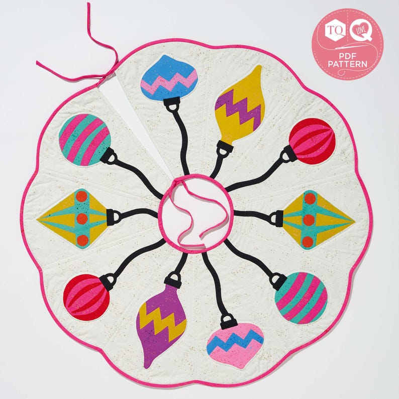 May include: A white fabric Christmas tree skirt with a pink scalloped edge. The skirt features eight colorful ornaments with different patterns and designs. The ornaments are attached to the center of the skirt with black strings.