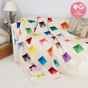 May include: A colorful quilt with a geometric pattern of diamonds in various shades of red, orange, yellow, green, blue, purple, and gray. The quilt is made of fabric with a subtle white polka dot pattern. The quilt is folded and draped over a white couch.