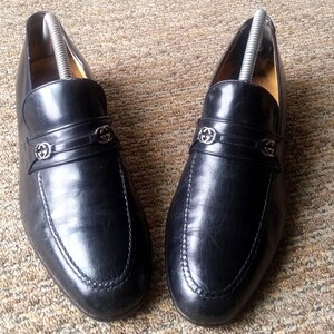 May include: A pair of black leather loafers with silver-tone hardware. The shoes feature a decorative strap across the vamp with a logo emblem. The soles have a stitched design. Shoe trees are inserted in the shoes.