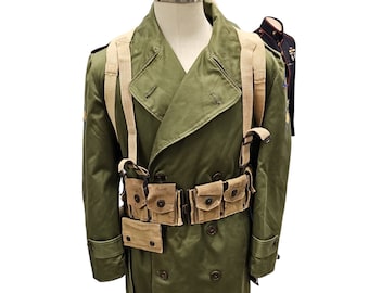 May include: An olive green military trench coat with a double-breasted design, displayed on a mannequin. The coat features a tan canvas belt and suspenders, along with multiple pouches. A second military jacket is visible in the background.
