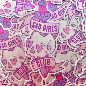 May include: A stack of stickers featuring a skull with hearts and a dagger with the text "Sad Girls Club". The stickers are pink and purple with a holographic finish.