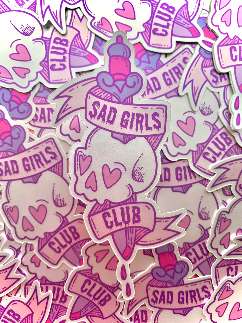 May include: A stack of stickers featuring a skull with hearts and a dagger with the text "Sad Girls Club". The stickers are pink and purple with a holographic finish.