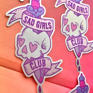May include: A silver sticker with a purple and pink design. The sticker features a skull with hearts, a dagger, and a banner that reads "Sad Girls Club".